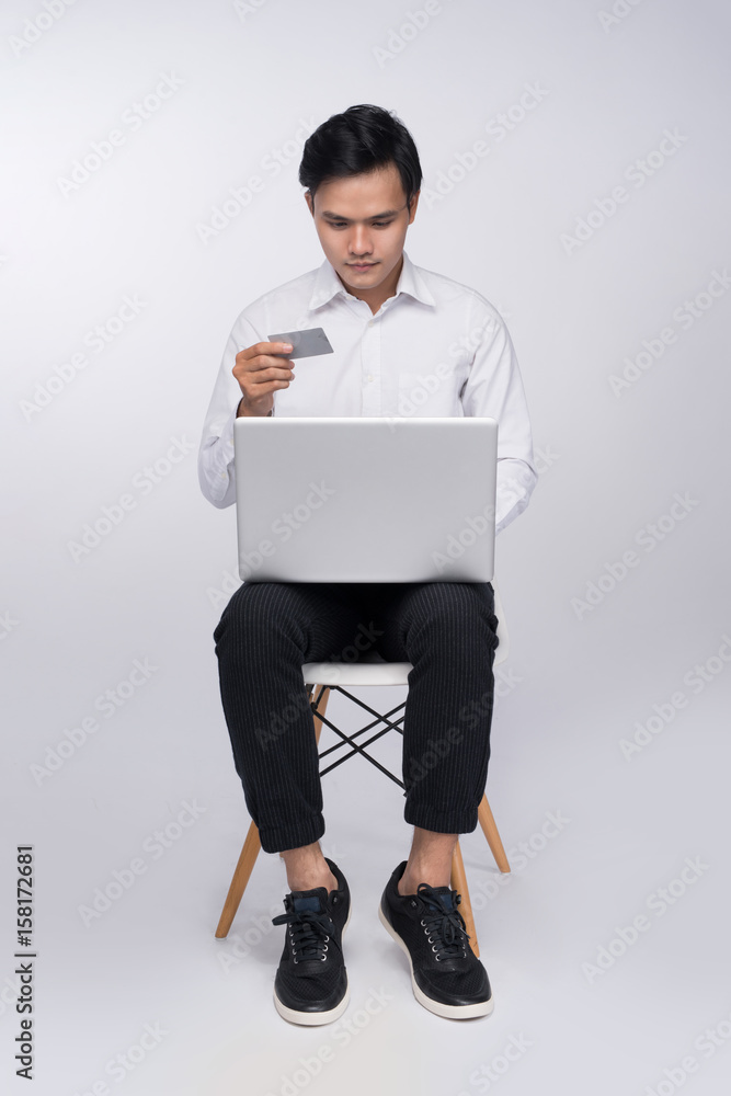 聪明休闲的亚洲男人坐在椅子上，用信用卡和笔记本电脑在线购物