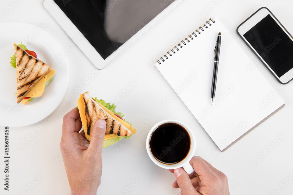Top view of person eating sandwiches and drinking coffee at workplace with electronics