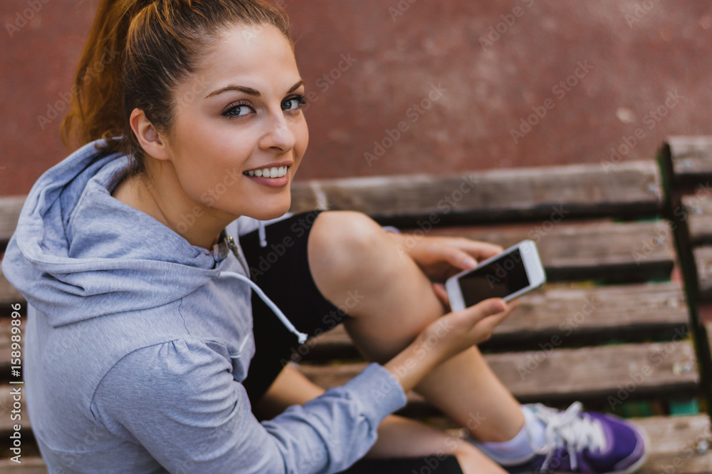 一位穿着运动装的漂亮年轻女子坐在公园的长椅上，手里拿着一部智能手机。