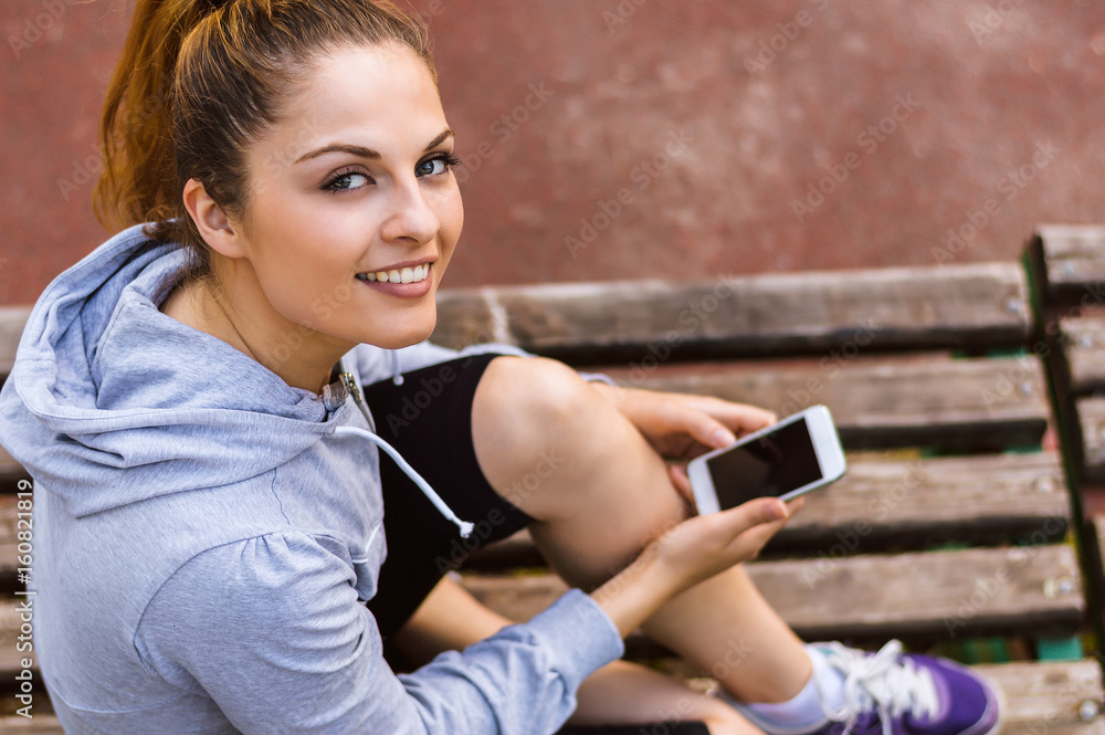Portrait of smiling girl using a smartphone while sitting on a wooden bench in a park.