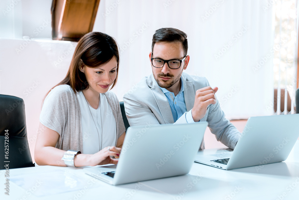 年轻的商人正在和他的女同事讨论，并在办公室里使用笔记本电脑。