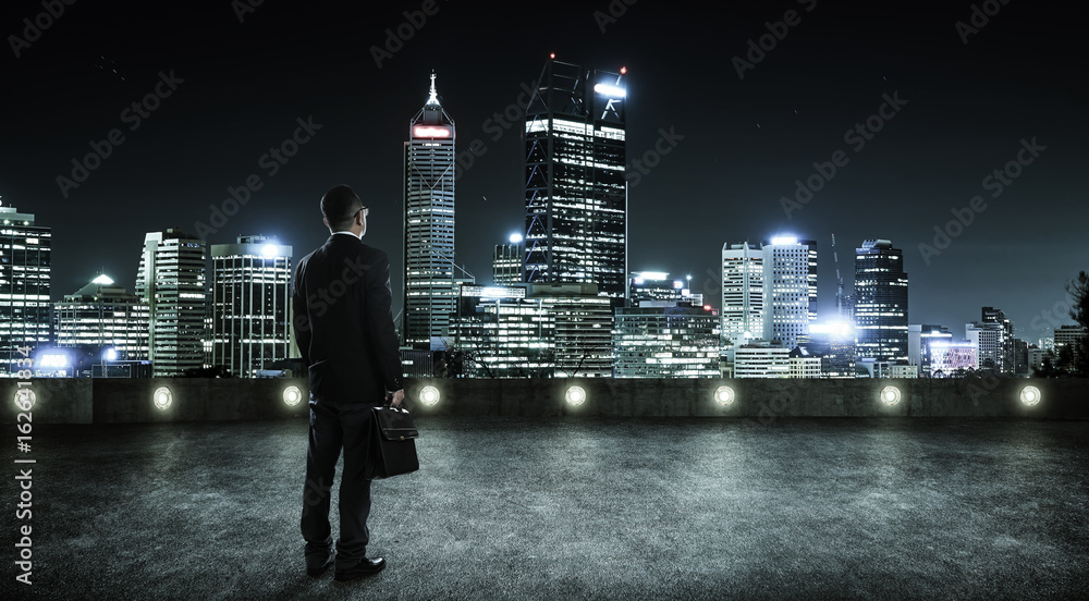 Businessman looking and thinking at roof top with modern city skyline background , night scene .