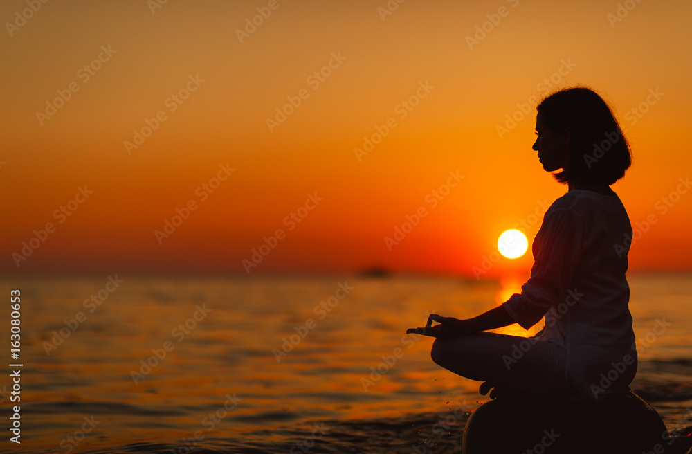 一位女士在日落海滩上练习瑜伽并以莲花姿势冥想。
