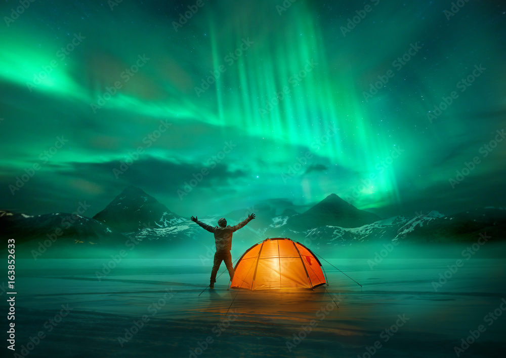 A man camping in wild northern mountains with an illuminated tent viewing a spectacular green northe