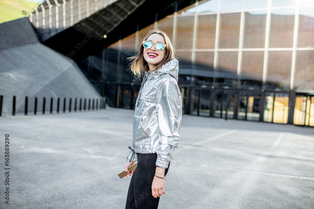 一位穿着银色夹克的时尚女性在现代建筑上走在户外的生活方式肖像