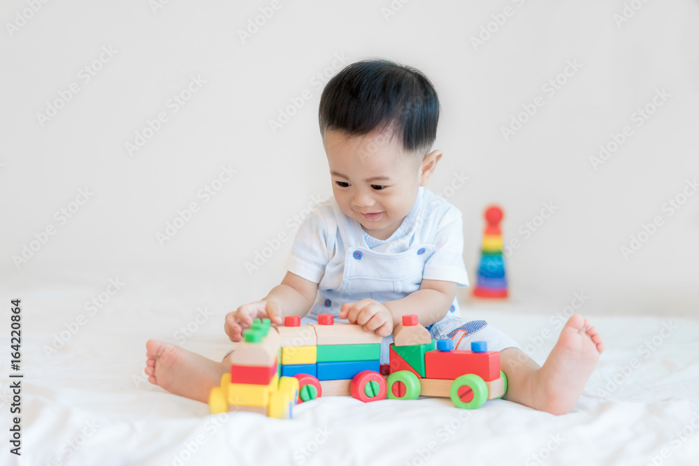 可爱的亚洲男婴9个月大，坐在床上，在家里玩彩色木制火车玩具。