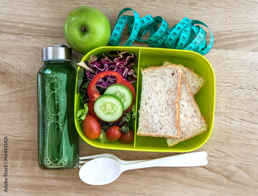 健康午餐盒，木质背景上有谷物面包、绿色蔬菜和果汁瓶