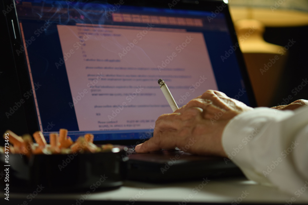 一名男子用手指夹着点燃的香烟，在笔记本电脑上工作