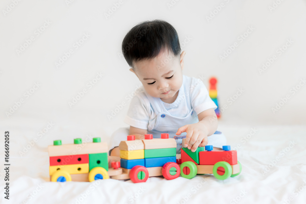 Adorable Asian baby boy 9 months sitting on bed and playing with color wooden train toys at home..