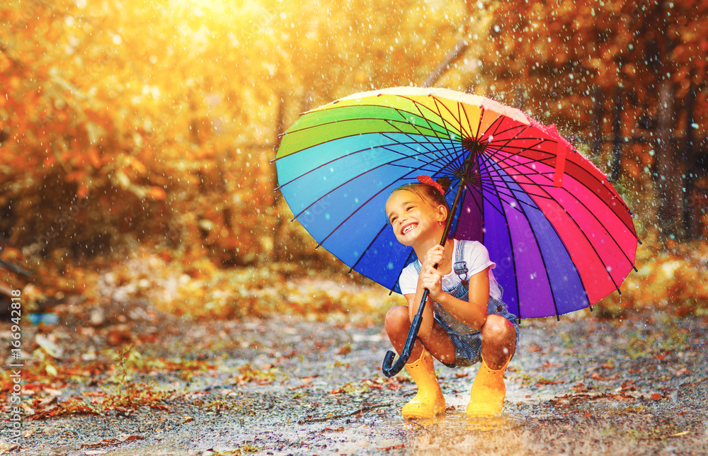 快乐有趣的小女孩，带着雨伞，穿着橡胶靴在水坑上跳跃。