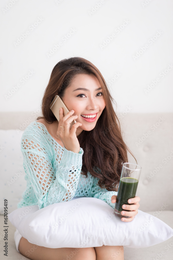 微笑的年轻亚洲女性用smartpho喝绿色新鲜蔬菜汁或冰沙