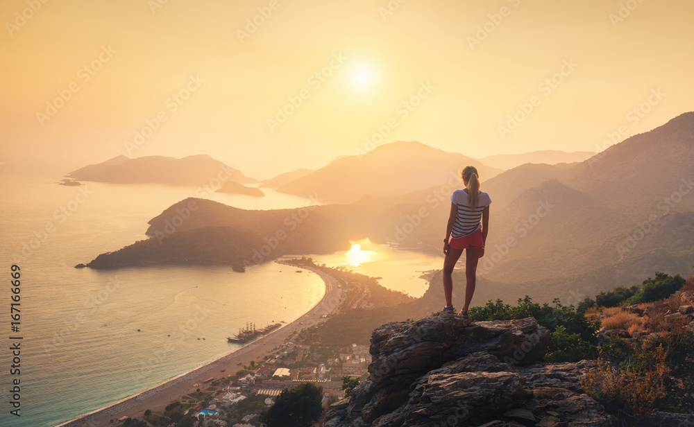 年轻的女人站在岩石顶部，看着五颜六色的日落中的海岸和山脉