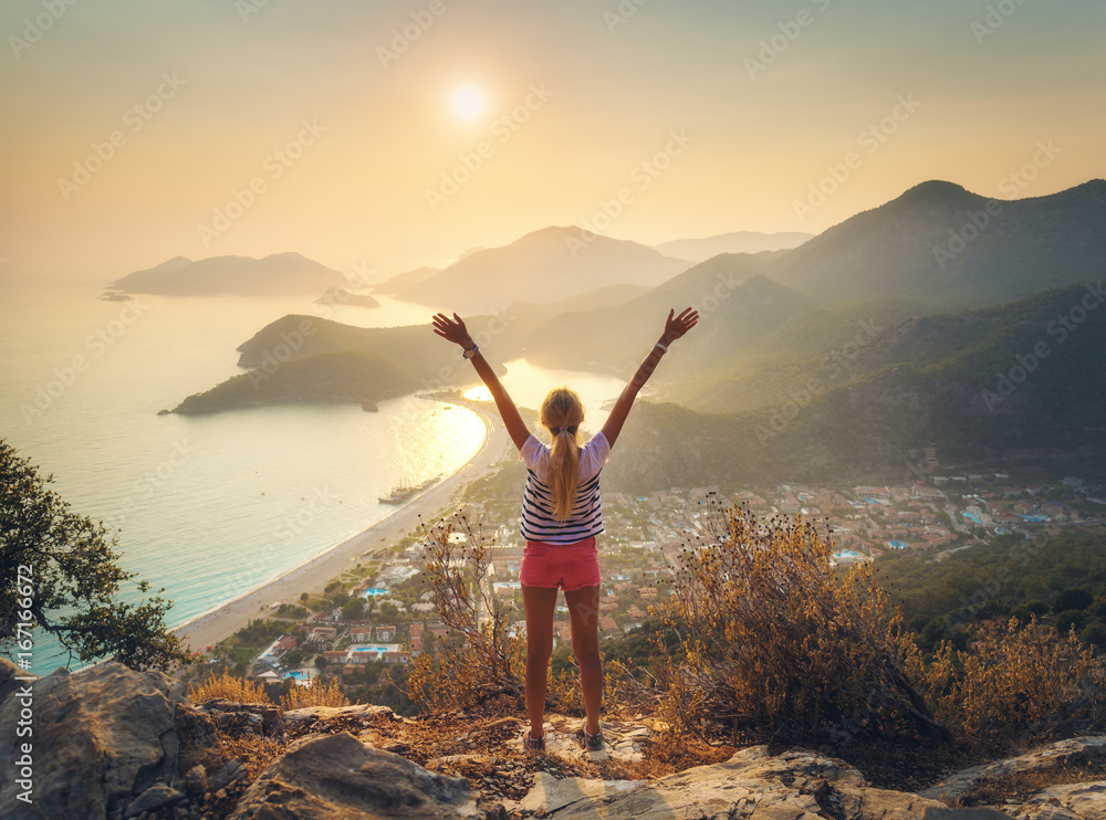 Happy woman with raised up arms standing on the top of rock and looking at the seashore and mountain