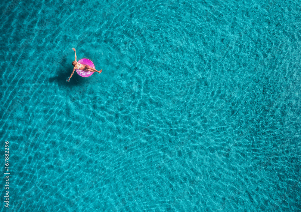 蓝色L的透明绿松石海中，年轻女子在粉色游泳圈上游泳的鸟瞰图