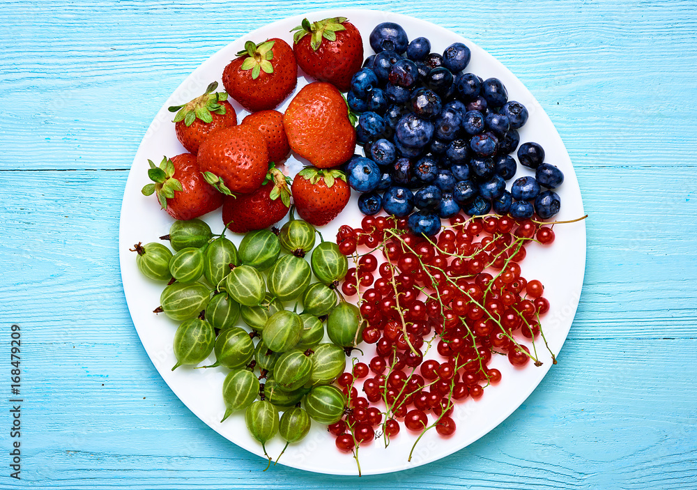 花园浆果放在白色盘子里。草莓、蓝莓、醋栗、醋栗放在蓝色木巴上