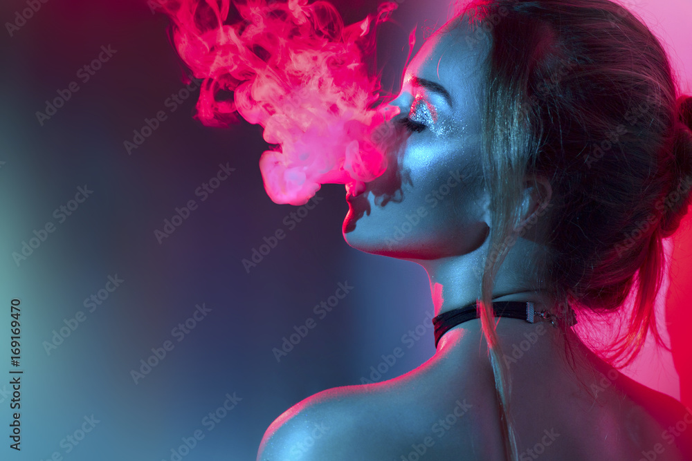 美丽模特的时尚艺术肖像，在明亮的灯光和五颜六色的烟雾中。吸烟的女孩