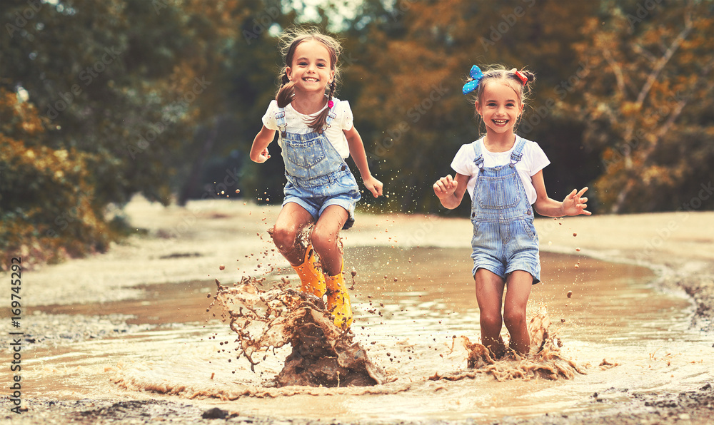 快乐有趣的姐妹双胞胎一个接一个地穿着橡胶靴在水坑上跳，笑得前仰后合。