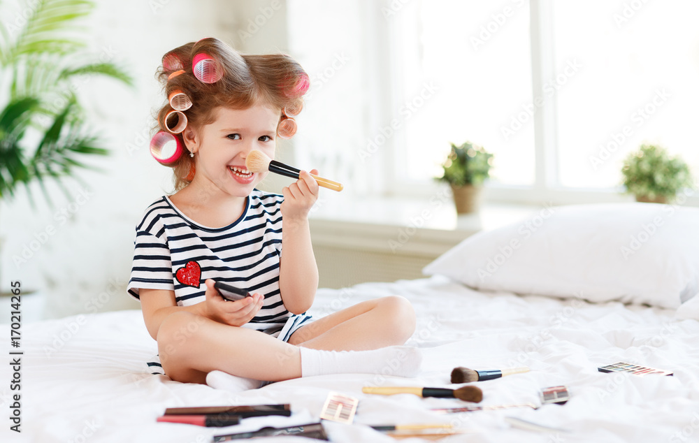美丽有趣的小女孩戴着卷发器在床上化妆大笑