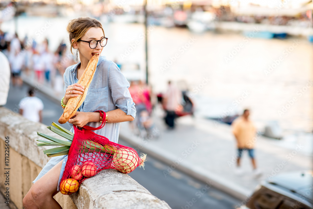 一位年轻女性的生活方式画像，她拿着装满新鲜食物的网袋，在餐厅里吃法棍面包