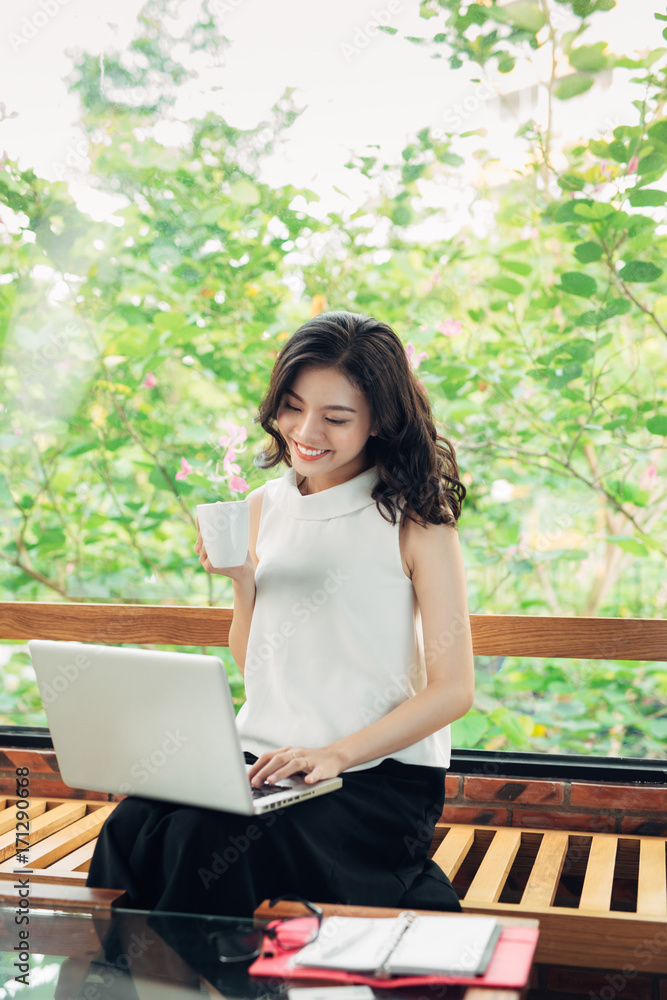 幸福的年轻商业女性的肖像，手里拿着马克杯，喝着咖啡，看着笔记本电脑。
