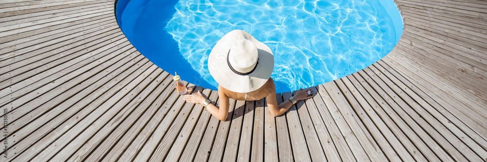 戴着大太阳帽，喝着鸡尾酒的女人在室外蓝水的圆形游泳池放松