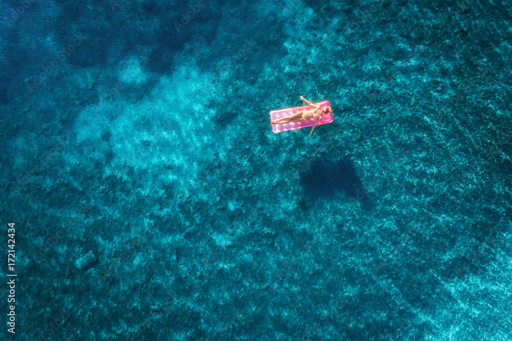 透明绿松石海中年轻女子在粉色充气床垫上游泳的鸟瞰图