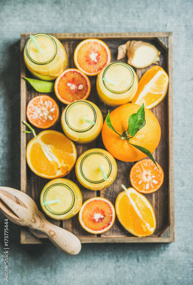 Healthy yellow smoothie with citrus fruit and ginger in bottles in wooden tray over grey concrete ba