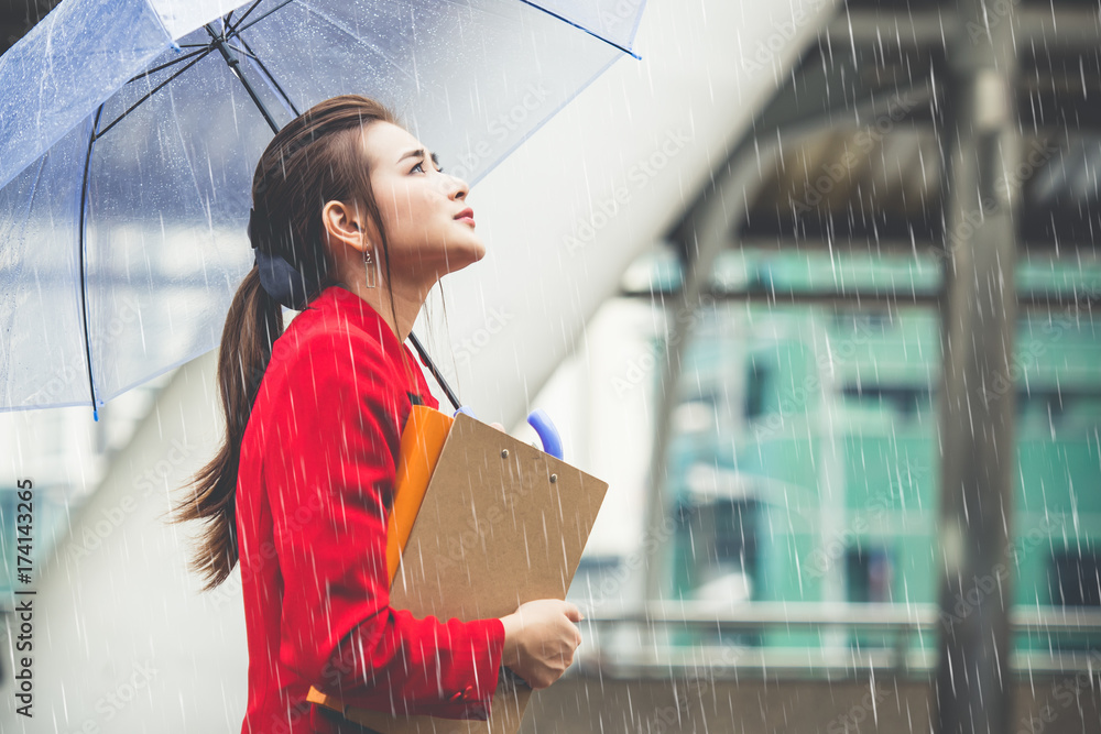 年轻的亚洲女商人拿着商务文件和雨伞在城市里雨中行走