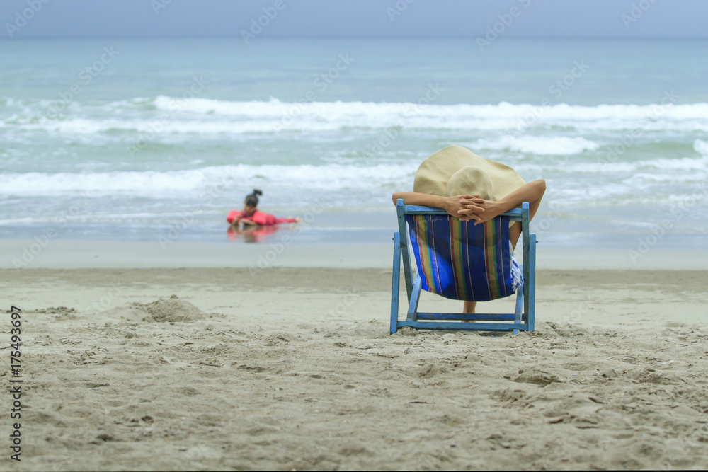 母亲坐在海滩的躺椅上，看着女儿玩耍，家庭生活方式旅行健康