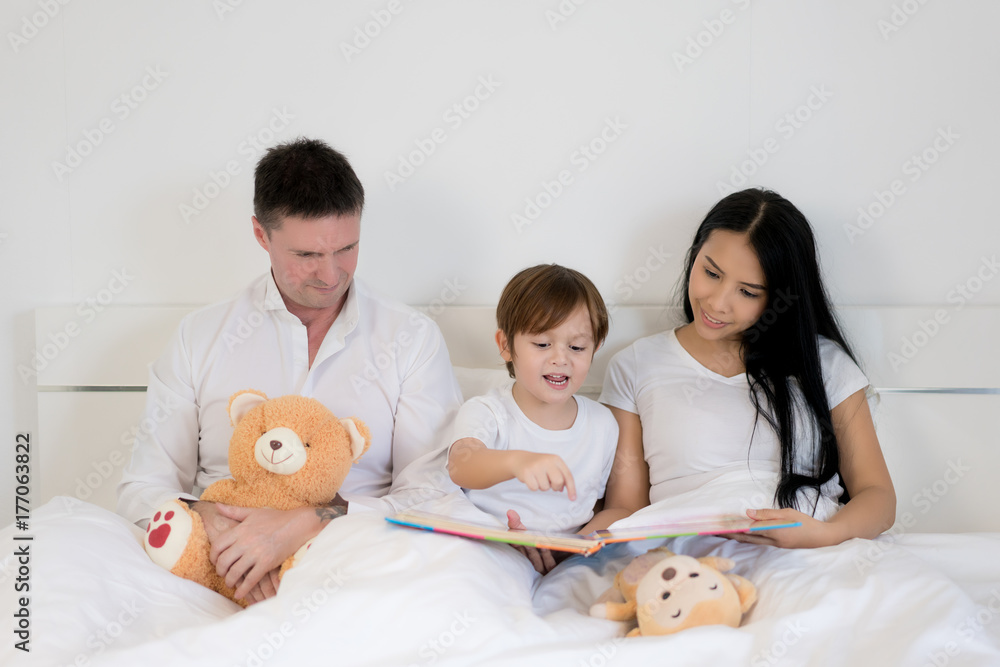 亚洲男孩与父亲和母亲一起在卧室的床上看书。幸福的家庭理念