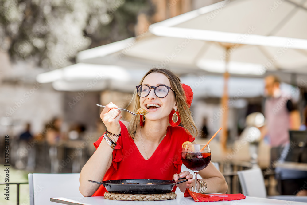一位身穿红色连衣裙的年轻女子坐在户外的餐厅里吃着传统的巴伦西亚米饭——海鲜饭