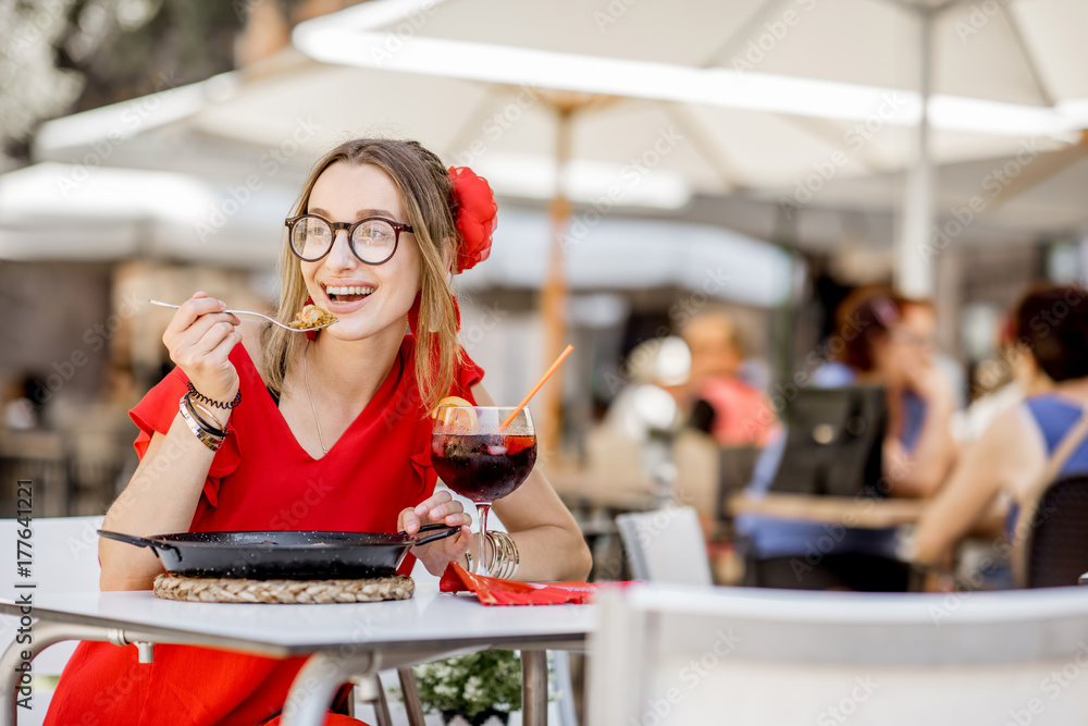 一位身穿红色连衣裙的年轻女子坐在户外的餐厅里吃着传统的巴伦西亚米饭——海鲜饭