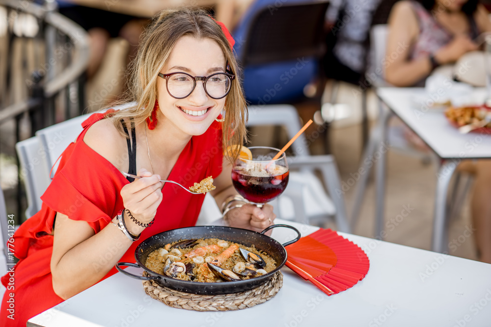 一位身穿红色连衣裙的年轻女子坐在户外吃着传统的巴伦西亚米饭——海鲜饭