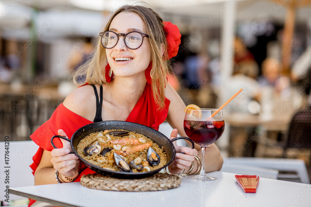 一位身穿红色连衣裙的年轻女士坐在户外吃着传统的巴伦西亚米饭——海鲜饭