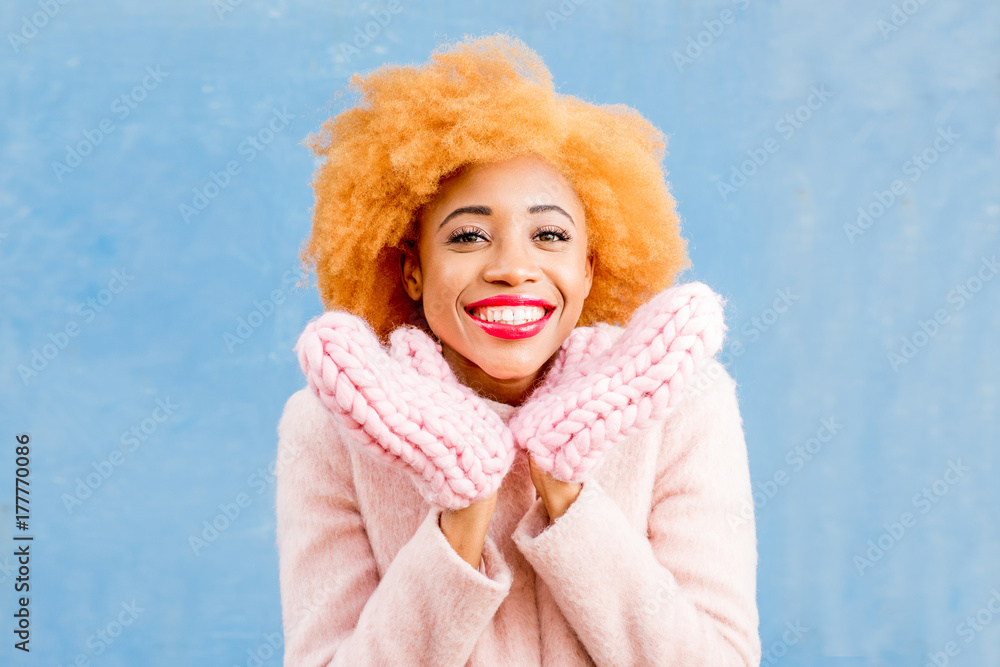 蓝色墙壁背景上一位身穿粉色外套、戴着针织手套的可爱非洲女性的肖像。Wi