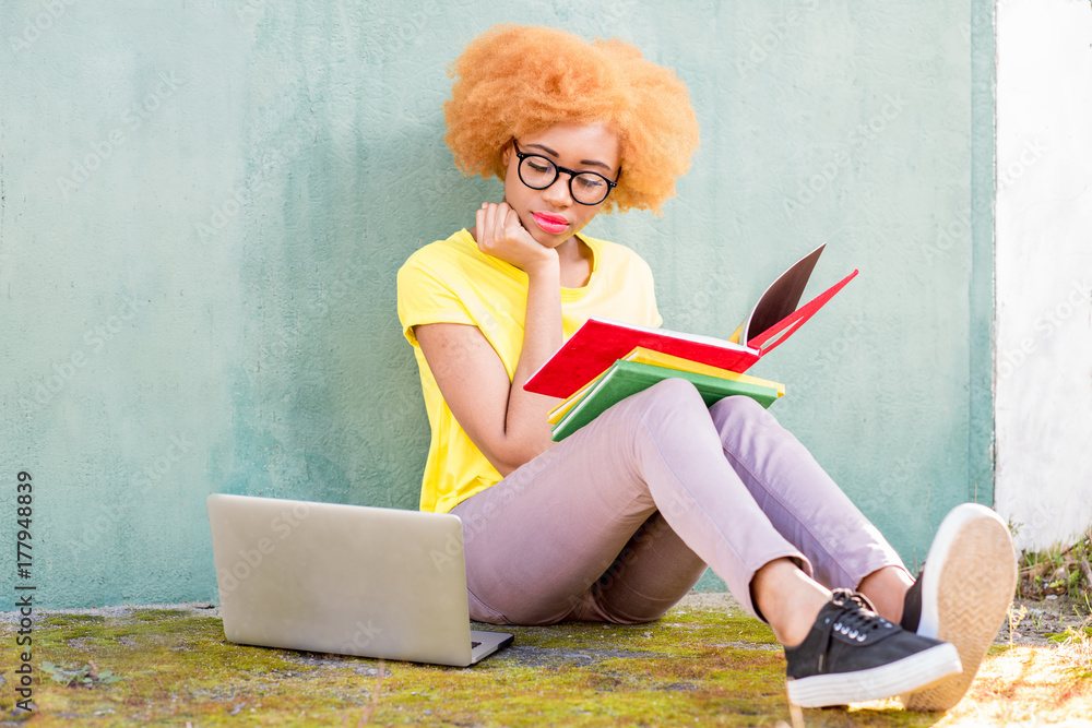 美丽的非洲女人，卷发，拿着书和笔记本电脑，坐在户外的草地上学习