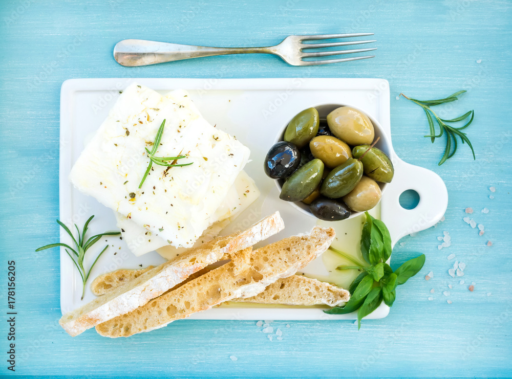 Fresh feta cheese with olives, basil, rosemary and bread slices on white ceramic serving board over 