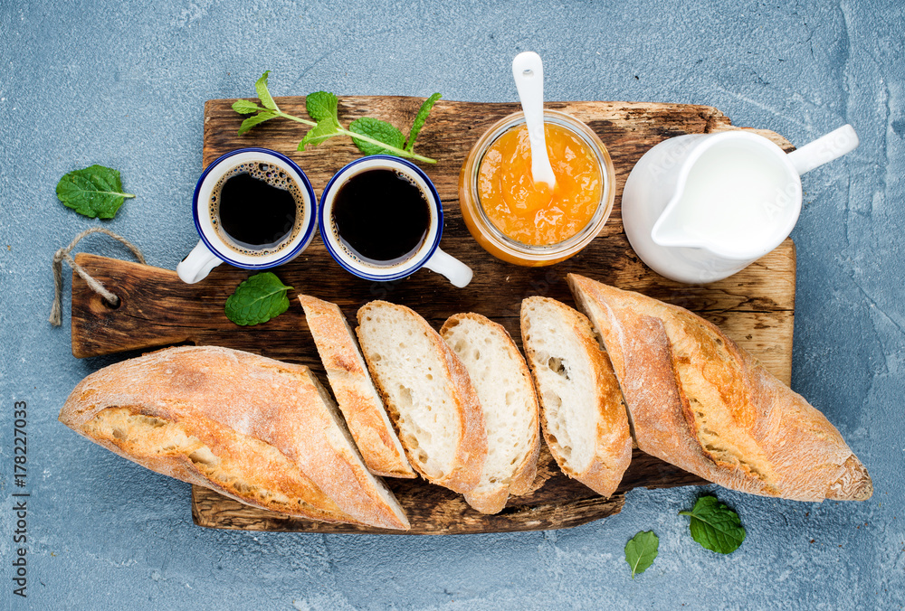 早餐套装。长棍面包、橙色果酱和咖啡装在乡村木板上的杯子里，上面是混凝土灰色的blu