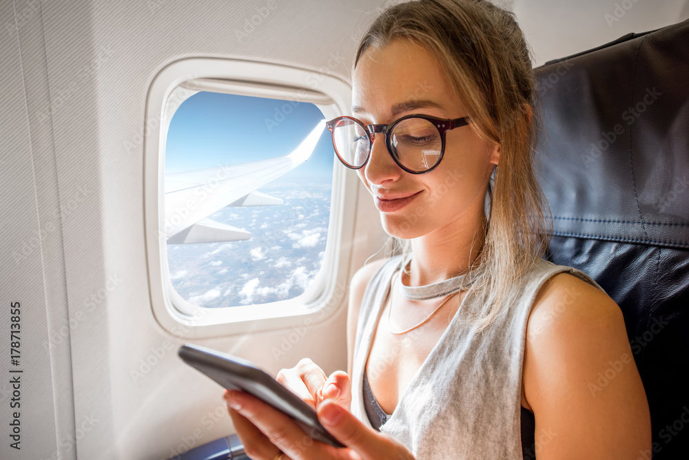 在空中飞行时，一名年轻女子拿着手机坐在靠近窗户的飞机座位上