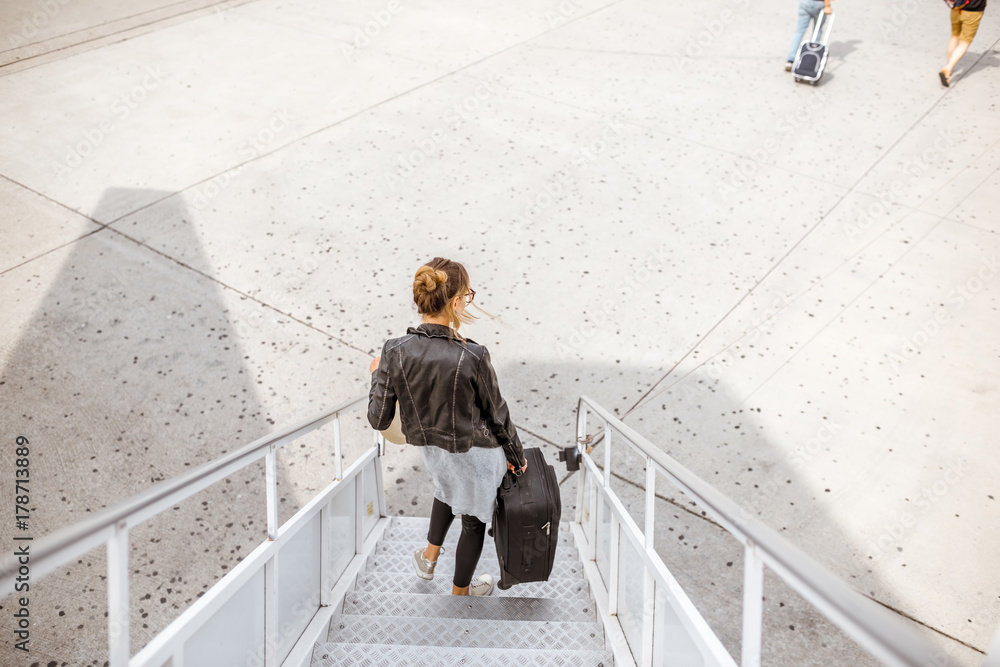 女子提着行李箱走下飞机楼梯，俯视图为地面背景。