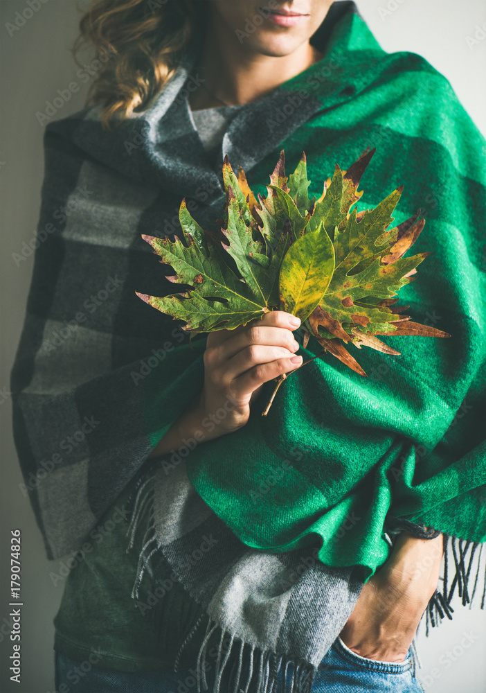 一个穿着温暖的羊毛绿色格子围巾或毯子和牛仔裤的女人，手里拿着秋天的落叶。