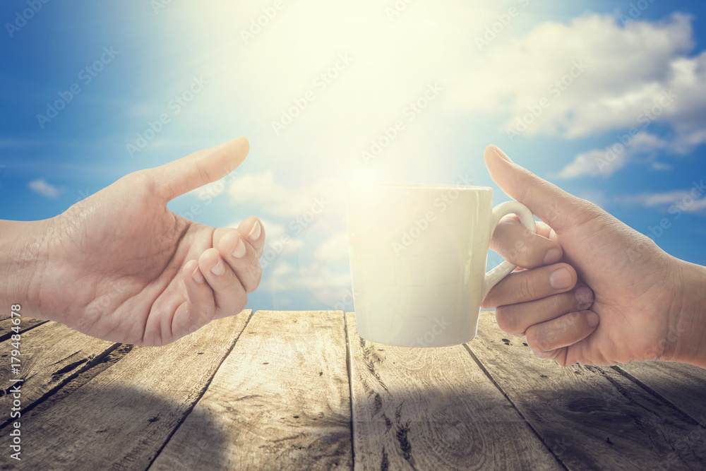 早上，在蓝天和阳光的木露台上，女人的手拿着一个白色的咖啡杯。好吧