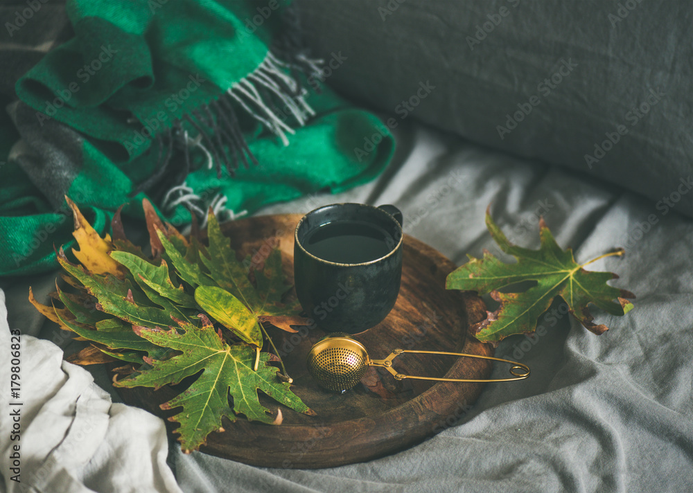 床上的秋季早茶。床单上的木托盘上放着一杯带筛子的红茶和落叶