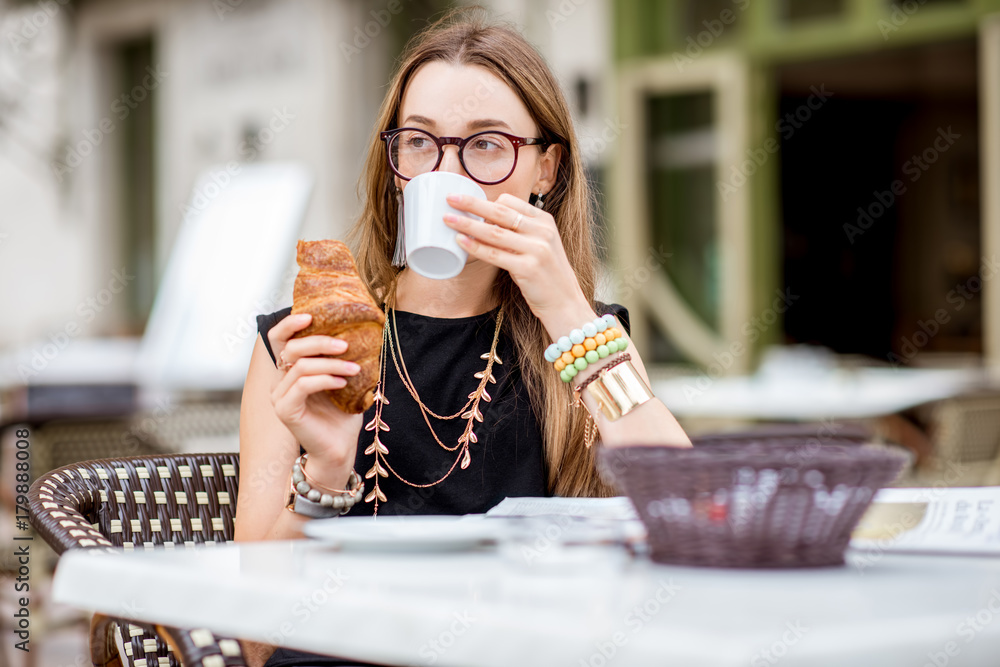 一位年轻女士坐在户外典型的法国咖啡馆吃早餐，配咖啡和羊角面包