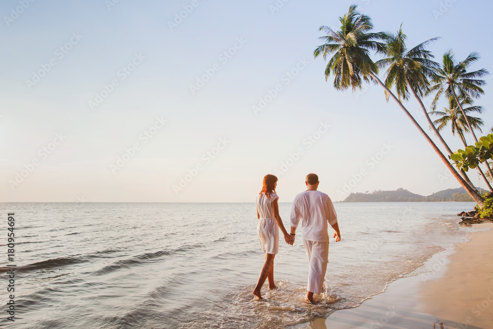 日落时分，浪漫的年轻情侣一起走在美丽的异国热带海滩上