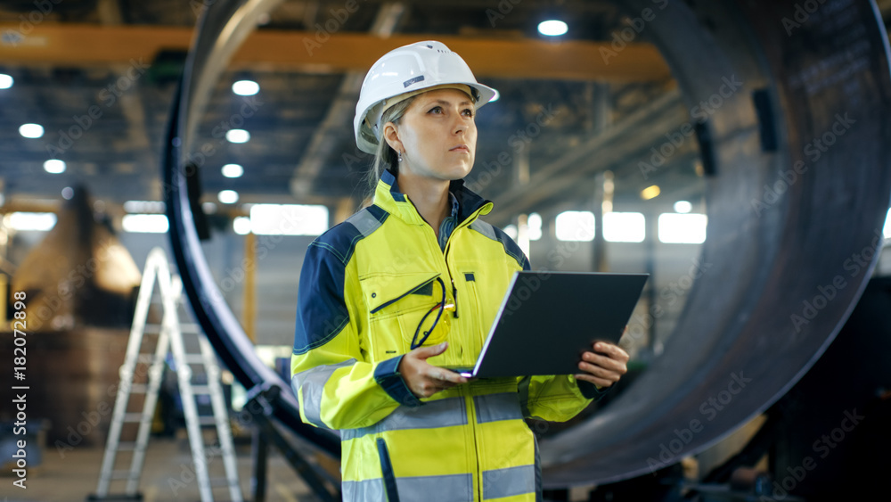 Female Industrial Engineer in the Hard Hat Uses Laptop Computer while Standing in the Heavy Industry