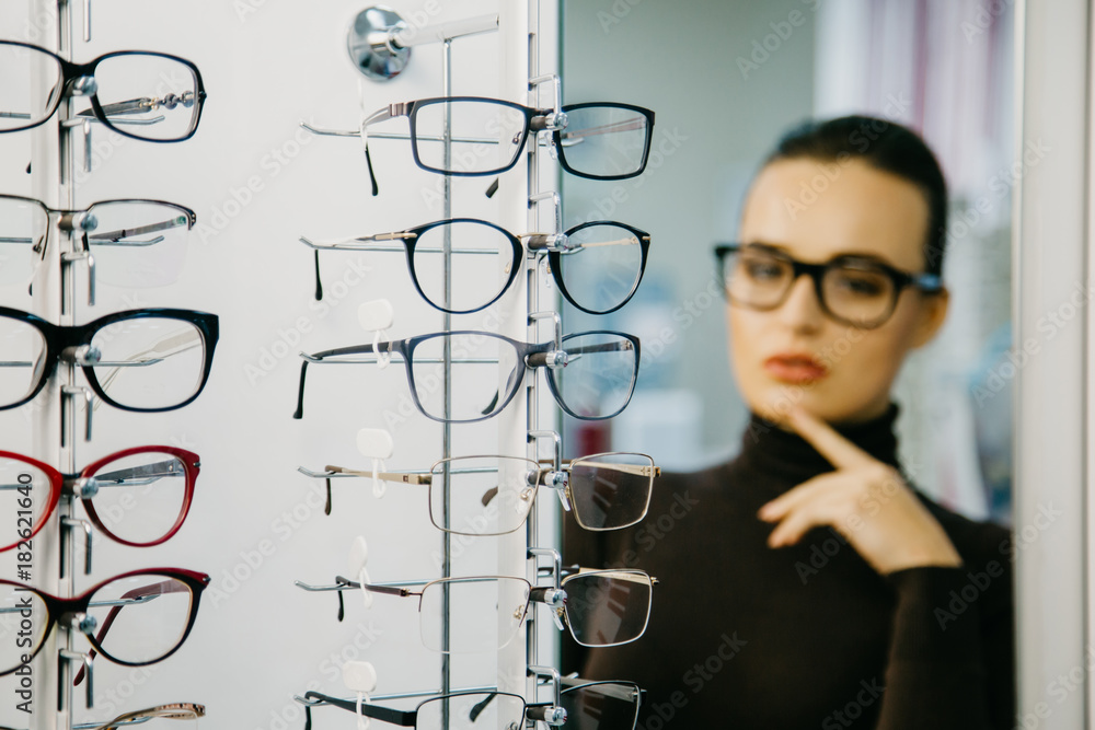 戴着眼镜站在光学商店里。背景上戴着眼镜的漂亮女孩。选择性聚焦