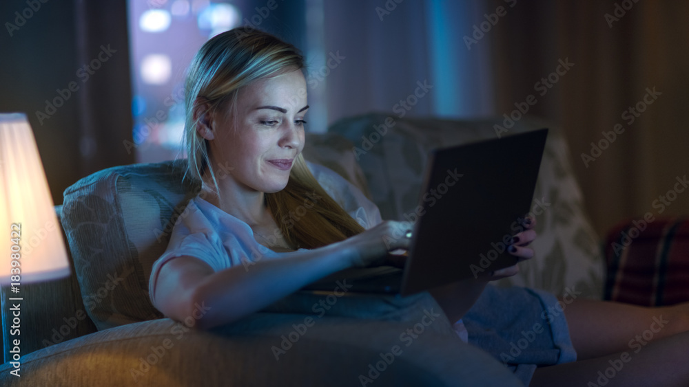 深夜美女躺在客厅的沙发上，在笔记本电脑上打字，面带微笑