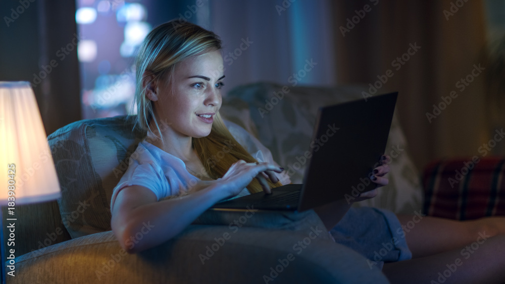 Late At Night Beautiful Woman Lying on Her Sofa in the Living Room, She Types on Her Laptop and Smil