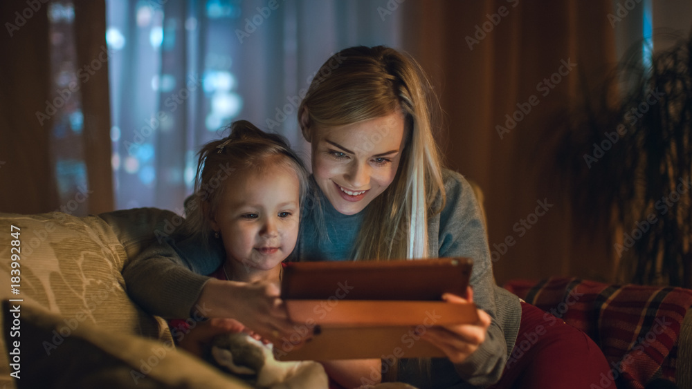 美丽的母亲和她的小女儿坐在客厅的沙发上，他们使用平板电脑C